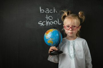 Fototapeta premium Portrait of little caucasian girl wearing glasses. Elementary school student holding the globe on the black school board background. Back to school concept
