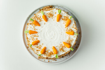 Delicious carrot cake decorated with mastic sweet carrots. Homemade carrot cake with yellow crumbs in the white plate on the white background. Flat lay