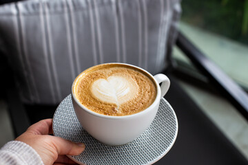 Woman is holding cup of hot cappuccino in cafe. Heart shape latte art for symbol of love.