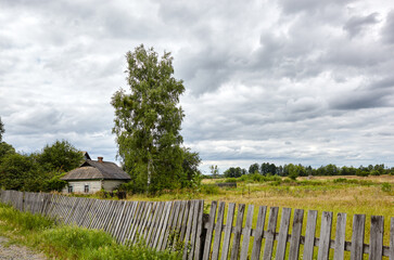 Beautiful summer rural landscape with wooden fence and old abadonted house. Meadow with trees and grass against the clouds sky
