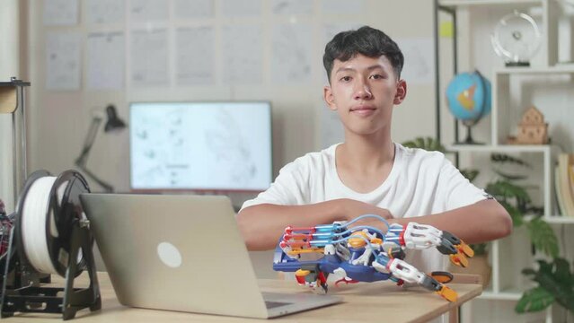 Asian Boy With A Laptop Smiling And Crossing His Arms To The Camera While Working About A Cyborg Hand At Home
