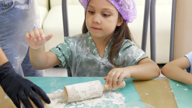 Pretty 5-year old girl in cap is going to roll out raw dough with a small rolling pin on a silicone mat at the master class. Preparing dough for baking pizza, pie. Children's activity about cooking.