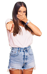 Fototapeta premium Young hispanic woman wearing casual white tshirt laughing at you, pointing finger to the camera with hand over mouth, shame expression