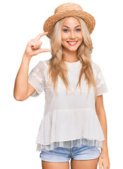 Obraz na płótnie Canvas Young blonde girl wearing summer hat smiling and confident gesturing with hand doing small size sign with fingers looking and the camera. measure concept.