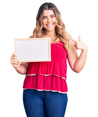 Young caucasian woman holding empty white chalkboard smiling happy pointing with hand and finger