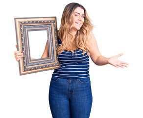 Young caucasian woman holding empty frame celebrating victory with happy smile and winner...