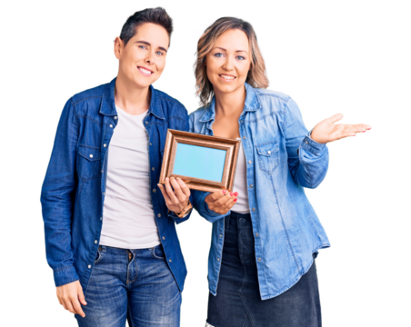 Couple of women holding empty frame smiling cheerful presenting and pointing with palm of hand looking at the camera.
