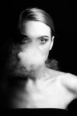 Fashion, style and make-up concept. Close-up black and white studio portrait of beautiful blonde woman blowing smoke from mouth. Model looking to camera with seductive look. Eyes in camera focus