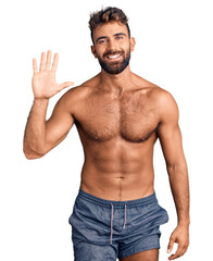 Young hispanic man wearing swimwear shirtless showing and pointing up with fingers number five while smiling confident and happy.