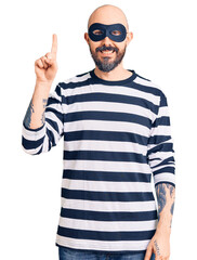 Young handsome man wearing burglar mask showing and pointing up with finger number one while smiling confident and happy.