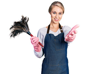 Beautiful young blonde woman wearing apron holding cleaning duster smiling friendly offering...