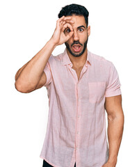 Hispanic man with beard wearing casual shirt doing ok gesture shocked with surprised face, eye looking through fingers. unbelieving expression.