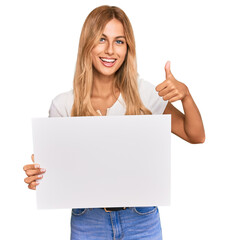 Beautiful blonde young woman holding blank empty banner smiling happy and positive, thumb up doing excellent and approval sign