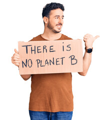 Young hispanic man holding there is no planet b banner pointing thumb up to the side smiling happy with open mouth