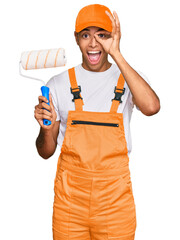 Young handsome african american man wearing cap and painter clothes holding painting roll smiling happy doing ok sign with hand on eye looking through fingers