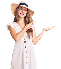Young beautiful blonde woman wearing summer dress and hat amazed and smiling to the camera while presenting with hand and pointing with finger.