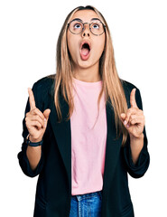 Hispanic young woman wearing business jacket and glasses amazed and surprised looking up and...
