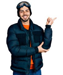 Young hispanic man with beard wearing snow wear and sky glasses with a big smile on face, pointing with hand and finger to the side looking at the camera.