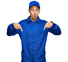 Bald man with beard wearing builder jumpsuit uniform pointing down with fingers showing advertisement, surprised face and open mouth