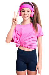 Obraz na płótnie Canvas Cute hispanic child girl wearing gym clothes and using headphones showing and pointing up with fingers number two while smiling confident and happy.