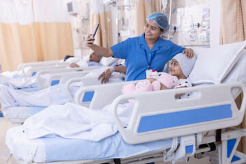 Fototapeta premium Female nurse taking selfie using mobile phone with girl patient sitting on bed at hospital 
