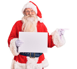 Old senior man with grey hair and long beard wearing santa claus costume holding banner smiling happy pointing with hand and finger to the side