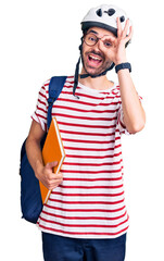 Young hispanic man wearing student backpack and bike helmet holding binder smiling happy doing ok sign with hand on eye looking through fingers
