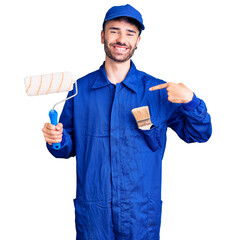 Young hispanic man wearing painter uniform holding roller pointing finger to one self smiling happy...