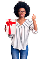 Young african american woman holding gift annoyed and frustrated shouting with anger, yelling crazy with anger and hand raised