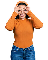 Young hispanic girl wearing casual white t shirt doing ok gesture like binoculars sticking tongue out, eyes looking through fingers. crazy expression.