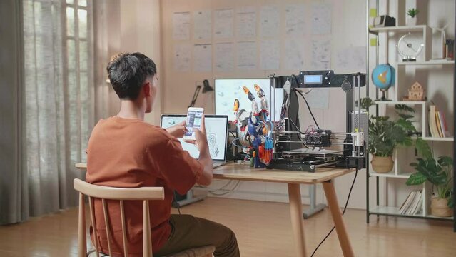 Back View Of A Boy With 3D Printing Looking At The Pictures On Smartphone While Designing A Cyborg Hand On A Laptop At Home
