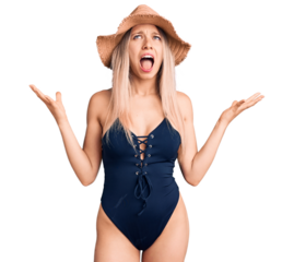 Young beautiful blonde woman wearing swimwear and summer hat crazy and mad shouting and yelling with aggressive expression and arms raised. frustration concept.