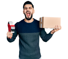 Young arab man with beard holding packing tape and cardboard box angry and mad screaming frustrated and furious, shouting with anger looking up.