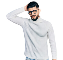 Young arab man with beard wearing elegant turtleneck sweater and glasses confuse and wondering about question. uncertain with doubt, thinking with hand on head. pensive concept.