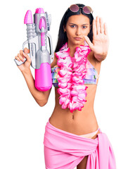 Young beautiful latin girl wearing bikini and hawaiian lei holding water gun with open hand doing...