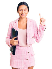 Young beautiful latin girl wearing business clothes holding binder surprised with an idea or question pointing finger with happy face, number one