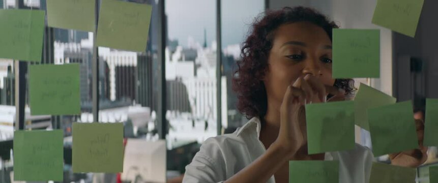 Young African American female employee looking at adhesive sticky notes on glass in the modern office, scrum agile methodology planning project. Shot through the glass