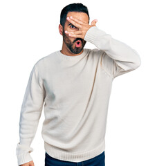 Young hispanic man with beard wearing casual white sweater peeking in shock covering face and eyes with hand, looking through fingers with embarrassed expression.