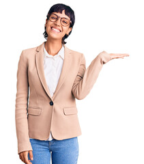 Young brunette woman with short hair wearing business jacket and glasses smiling cheerful presenting and pointing with palm of hand looking at the camera.