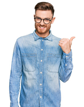 Young redhead man wearing casual denim shirt smiling with happy face looking and pointing to the side with thumb up.