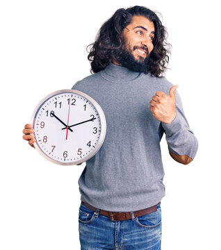 Young arab man holding big clock pointing thumb up to the side smiling happy with open mouth