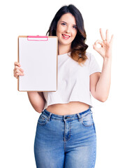Beautiful young brunette woman holding clipboard with blank space doing ok sign with fingers, smiling friendly gesturing excellent symbol