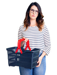 Beautiful young brunette woman holding supermarket shopping basket thinking attitude and sober expression looking self confident