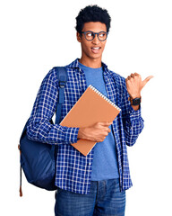 Young african american man wearing student backpack holding book smiling with happy face looking and pointing to the side with thumb up.