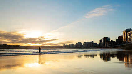 Fototapeta premium Beautiful panoramic view of the sunrise over the bay of San Lorenzo in Gijón, Spain. Sunrise over Gijón with reflections of the sun on the wet sand. Backlit silhouette in Gijón
