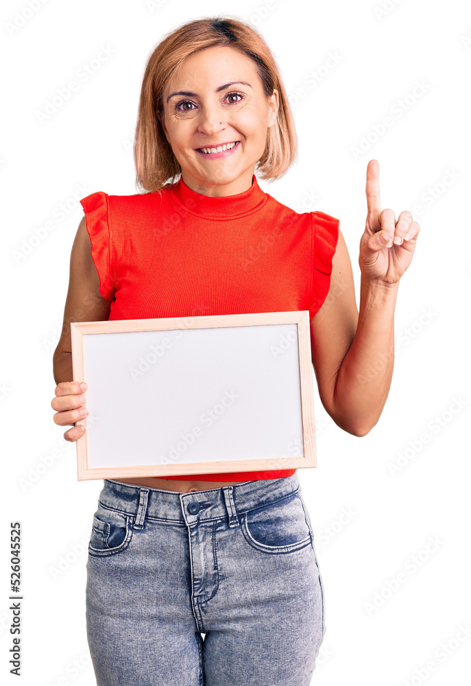 Poster Young blonde woman holding empty white chalkboard surprised with an idea or question pointing finger with happy face, number one