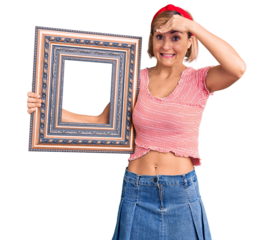 Young blonde woman holding empty frame stressed and frustrated with hand on head, surprised and angry face