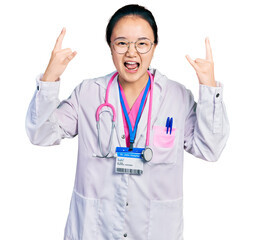 Young chinese woman wearing doctor uniform and stethoscope shouting with crazy expression doing rock symbol with hands up. music star. heavy music concept.