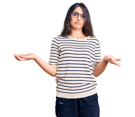 Brunette teenager girl wearing casual clothes and glasses clueless and confused expression with arms and hands raised. doubt concept.