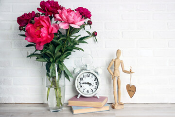 Purple peony flowers bouquet, books, alarm clock and wooden dummy on the white brick wall.